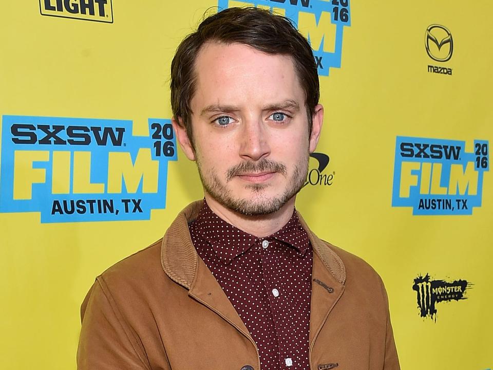 elijah wood wearing a brown blazer and standing in front of a yellow background