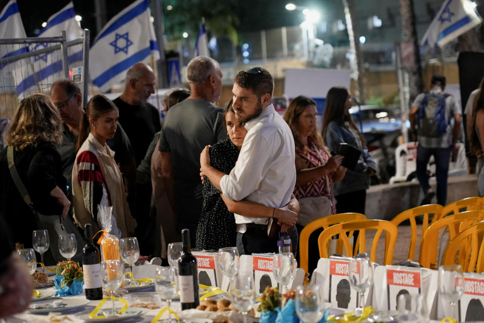 An couple embraces among others near Israeli flags by a table set with placeholders that read: Hostage.