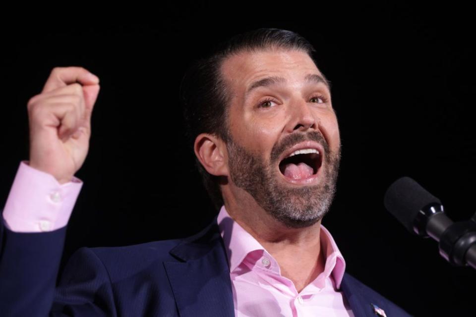 donald trump jr wearing a dark blue suit jacket and pink shirt, standing in front of a microphone and raising his right hand