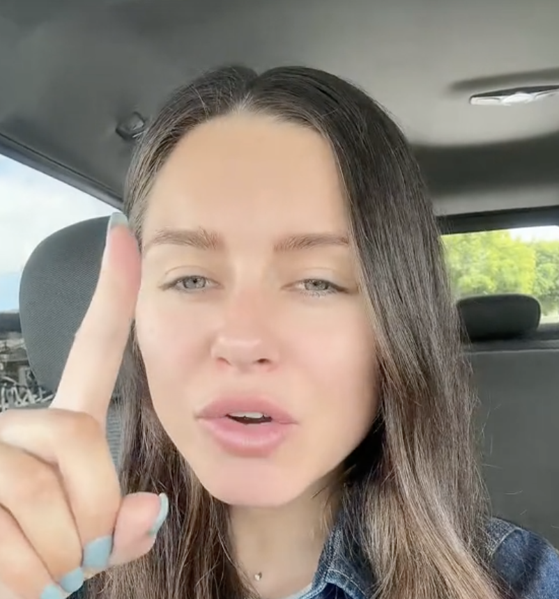 Woman holding up her index finger, speaking in a car, looking into the camera