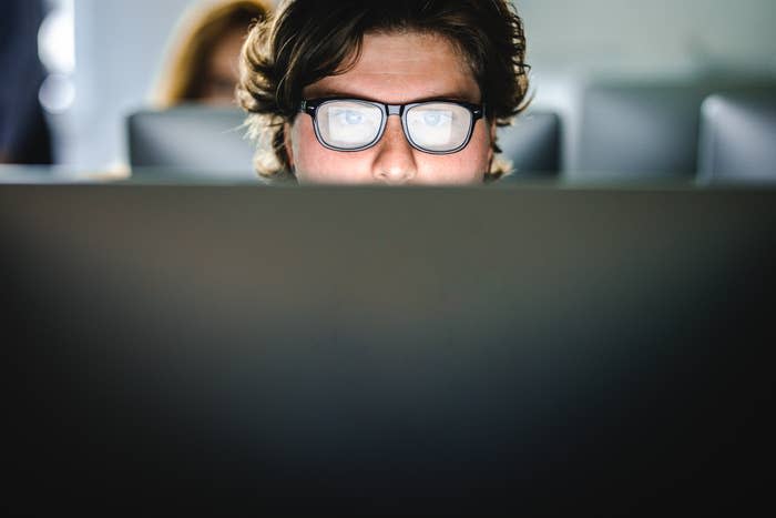 A person with glasses is closely looking at a computer screen. Only the top part of the head and eyes are visible. Another individual is blurred in the background