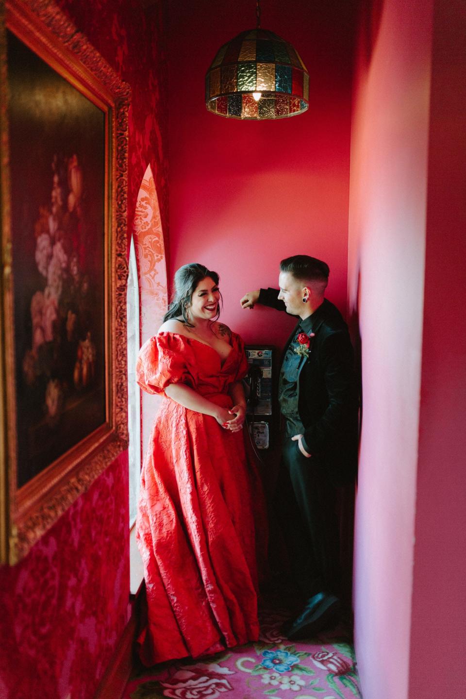 A man in a black suit and a woman in a red dress look at each other in a red hallway.