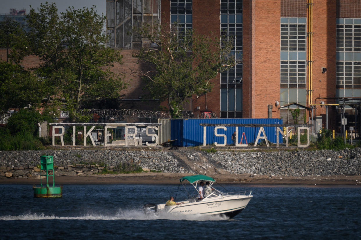 Rikers Island sign seen from the water with the building behind and a motorboat cruising by. 