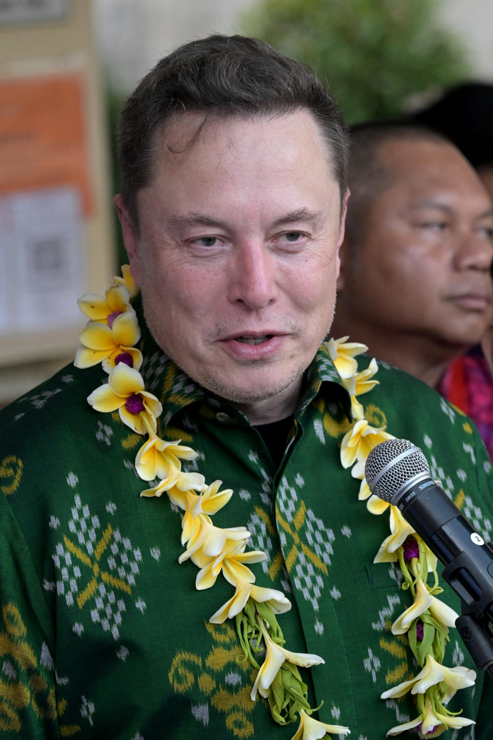 Elon Musk is speaking at the event wearing the traditional green patterned shirt and a yellow lei around his neck, with another person standing behind him.