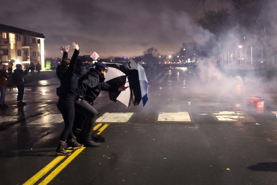 A small group of people hold open umbrellas against a cloud of smoke in a street at night.