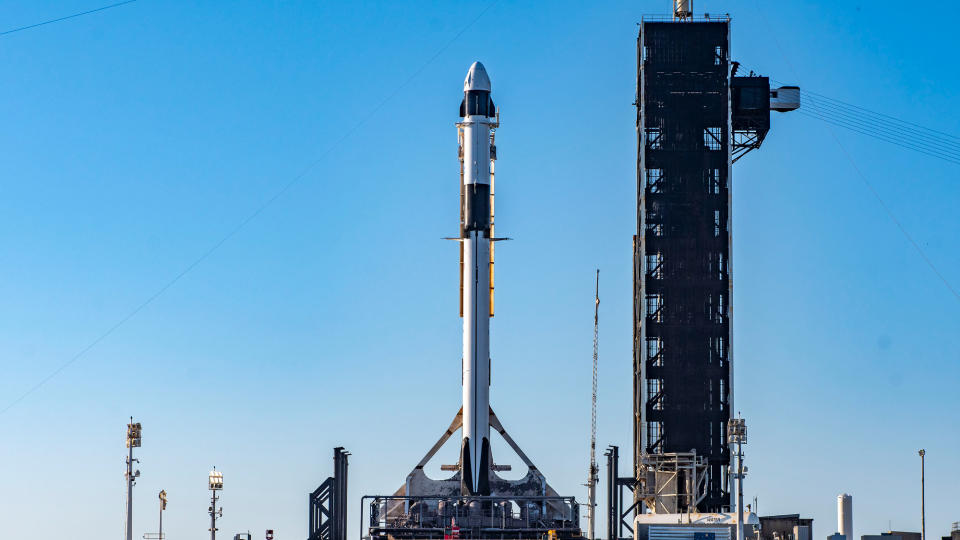 a white and black rocket topped with a white capsule stand on a launch pad with a blue sky in the background.