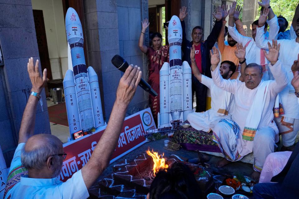 men in white clothing hold hands up in prayer sitting cross legged in front of cutouts of a white rocket