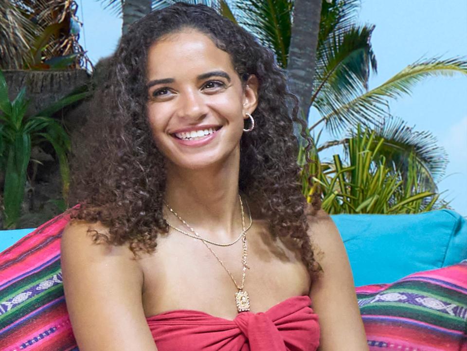 Olivia Lewis sits on a beach chair in a red top and jumpsuit while smiling at the camera.