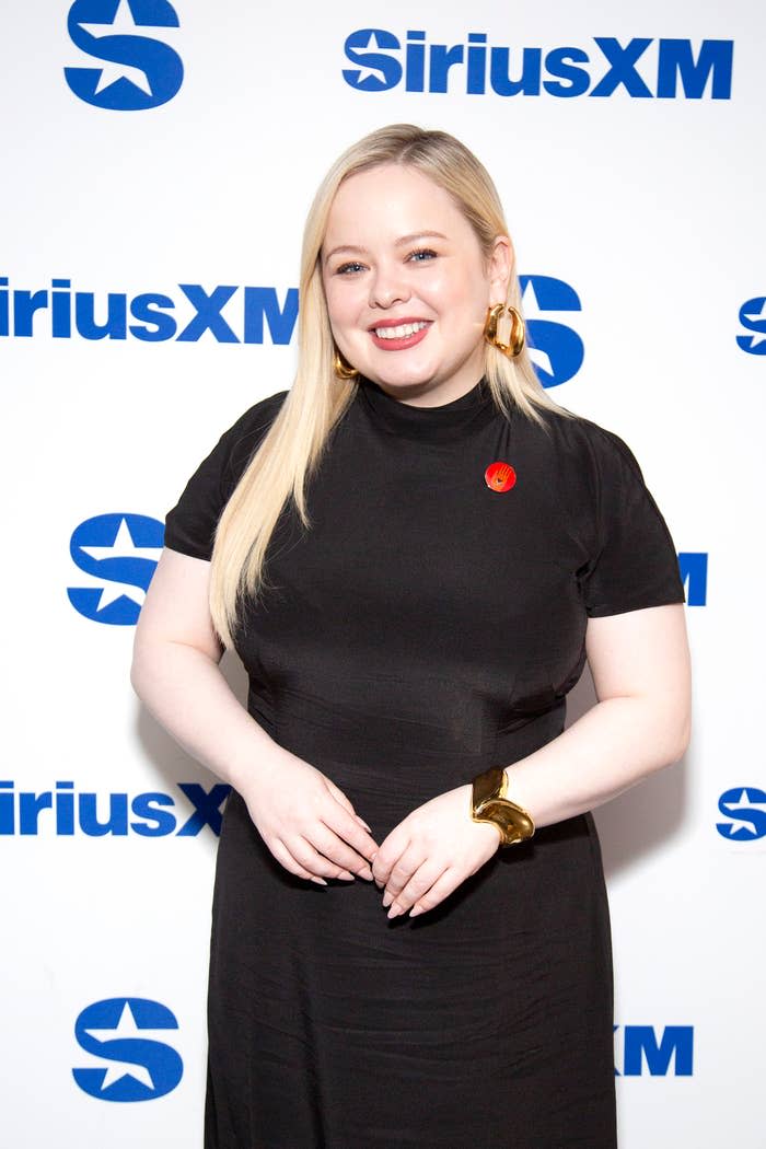 Nicola Coughlan smiles while posing in a dress with a pin and accessories at a SiriusXM event