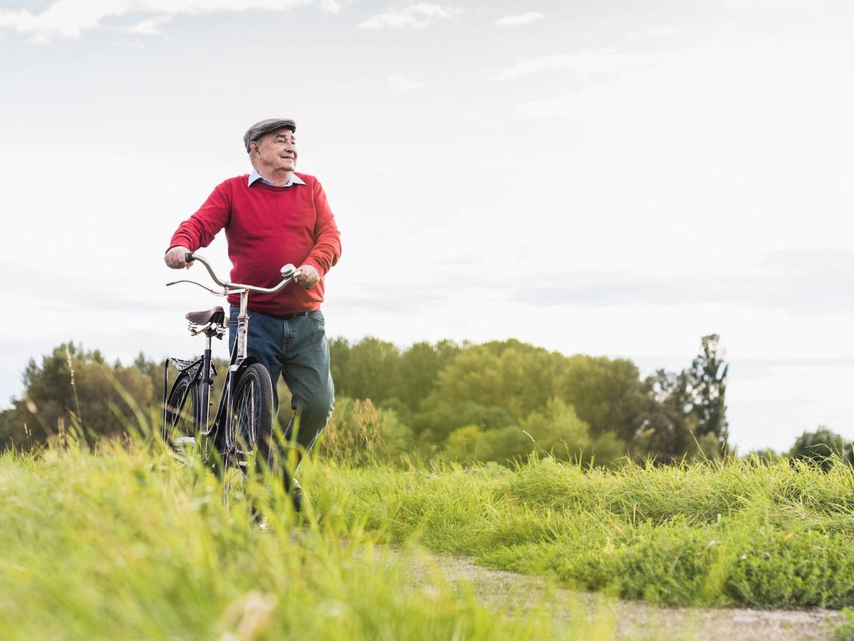 An older man in a sweater and flatcap walking a bike through a path with greenery either side.