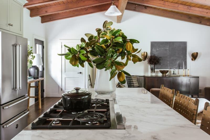 Large kitchen island, white marble countertop with gray veining, stove set in to island, large woven back bar stool, large plant on countertop
