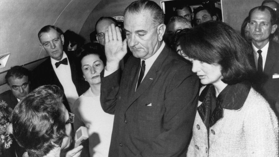 lyndon b johnson raises one hand to take an oath while standing next to jacqueline kennedy, wife lady bird johnson, and several other men