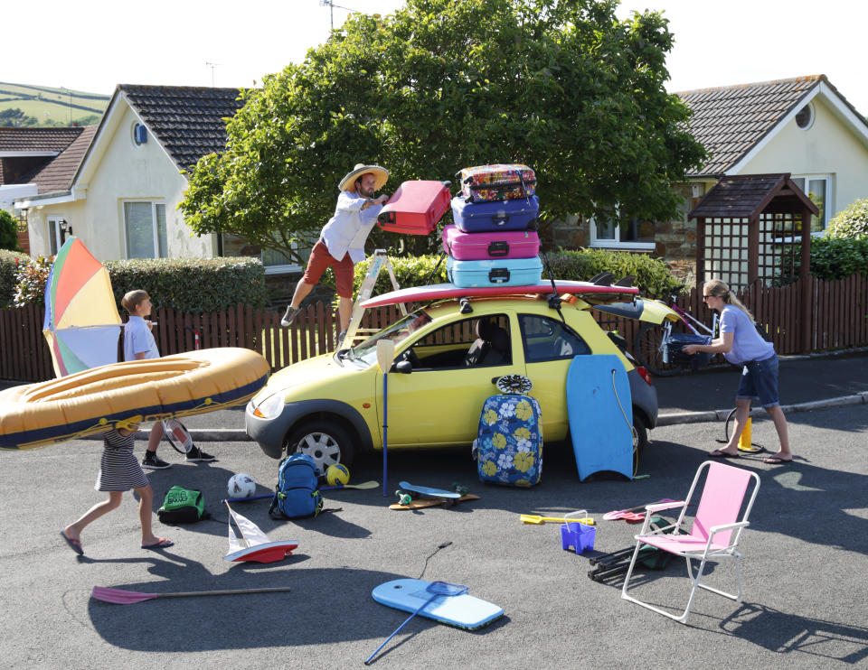 A family trying to fit all their suitcases, bookie boards and camp chairs into a tiny yellow car.