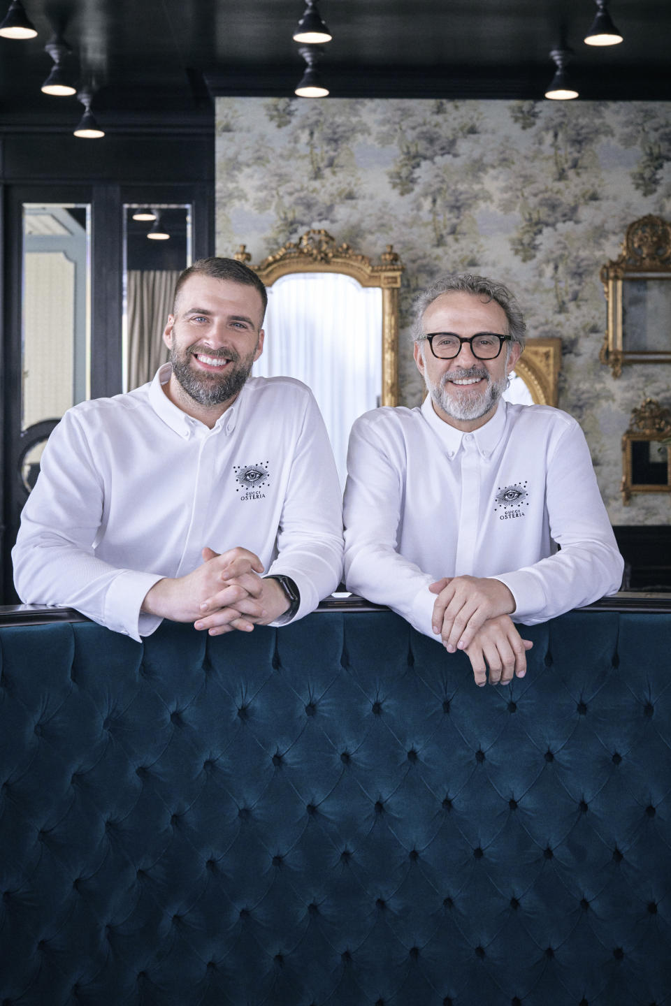 Antonio Iacoviello and Massimo Bottura pose smiling standing behind a blue quilted velvet couch, their arms resting on it.