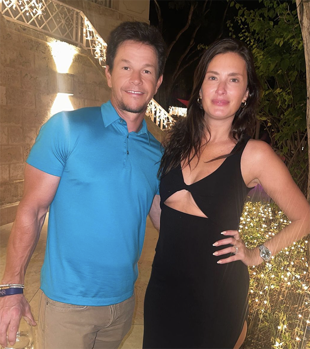 Mark Wahlberg in a blue shirt smiles next to his wife Rhea Durman in a black dress with a cutout on the chest and one hand on her hip.