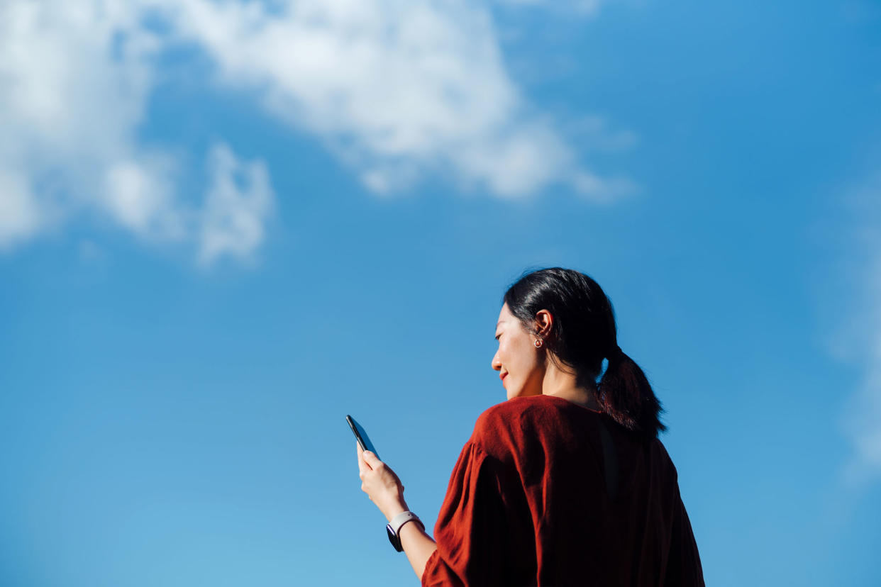 Low angle portrait of young woman using smartphone against blue sky