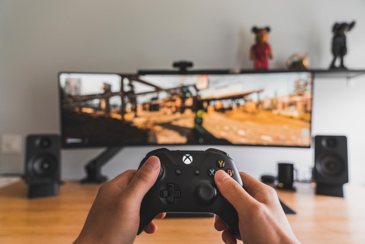  A person holding an Xbox controller in the foreground. The background, which is blurred, shows a game on a wide-screen TV. 