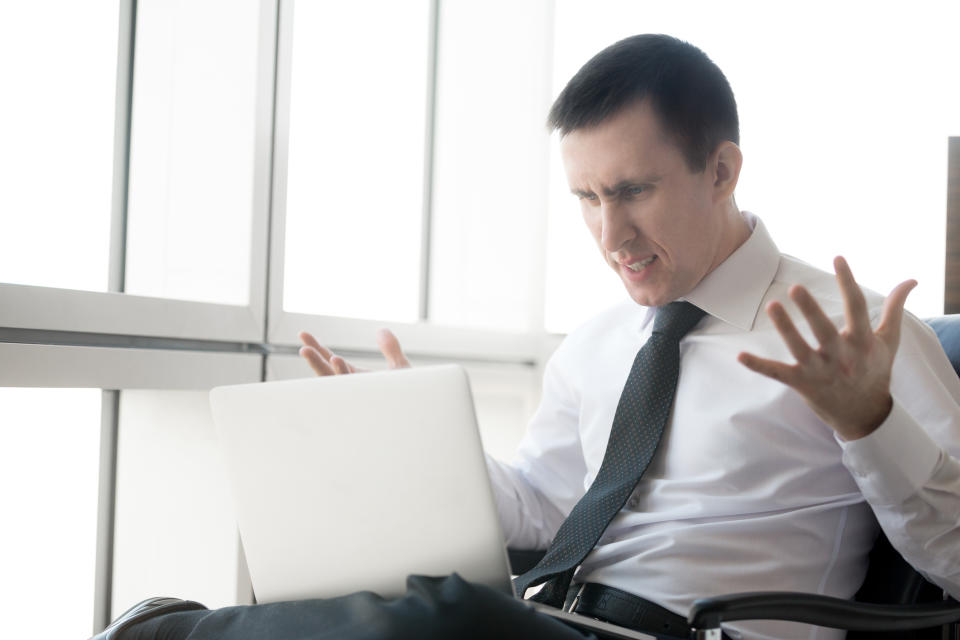 A visibly frustrated businessman throwing his hands up in the air while reading information on his laptop.