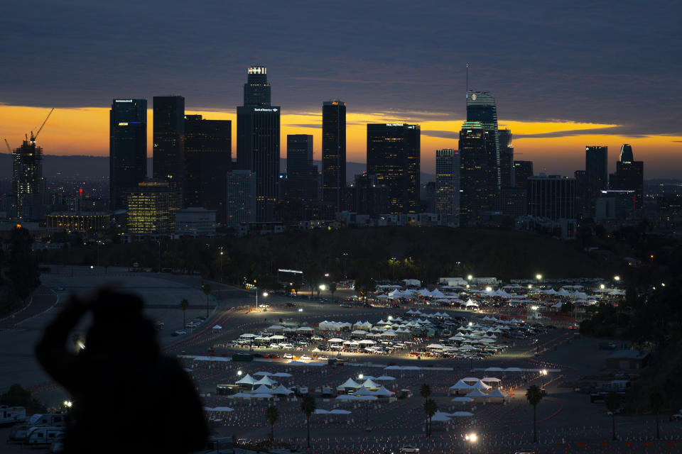 FILE - In this Jan. 27, 2021, file photo, a spectator stands on a hilltop overlooking downtown Los Angeles as motorists line up for COVID-19 vaccinations and testing in the parking lot of Dodger Stadium in Los Angeles. One of the largest vaccination sites in the country temporarily shut down Saturday because dozen of protesters blocked the entrance, stalling hundreds of motorists who had been waiting in line for hours, the Los Angeles Times reported. The Los Angeles Fire Department shut the entrance to the vaccination center at Dodger Stadium about 2 p.m. as a precaution, officials told the newspaper. (AP Photo/Jae C. Hong, File)