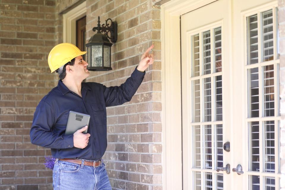 Latin descent man inspects home exterior. He could be a home inspector, insurance adjuster, exterminator or a variety of blue collar occupations. He holds a digital tablet, wears hard hat and blue work shirt.