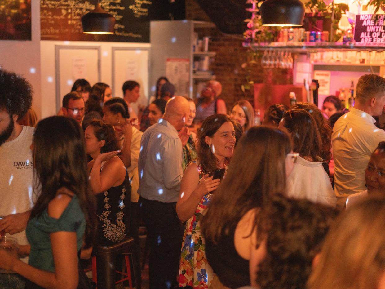 a crowd of adults at henrietta hudson chatting and and drinking assorted cocktails and beers. there are bright party lights and floral decorations