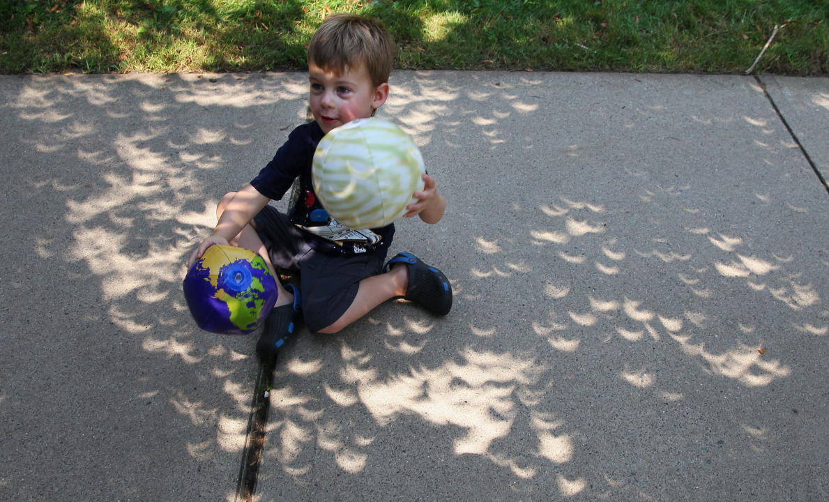 Elliot Braun aligns his toy Earth and Sun as crescent shaped beams of light from the partial eclipse shining through foliage create images of the sun on the sidewalk around him as hundreds of people gathered at Western University to view the partial solar eclipse, in London, Ont. on Monday, Aug. 21, 2017. THE CANADIAN PRESS/Dave Chidley