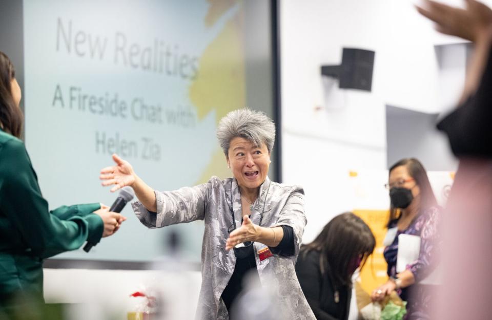 A gray-haired woman in a gray jacket, her hands extended, looks to the side. Next to her, a woman wearing green holds a mic