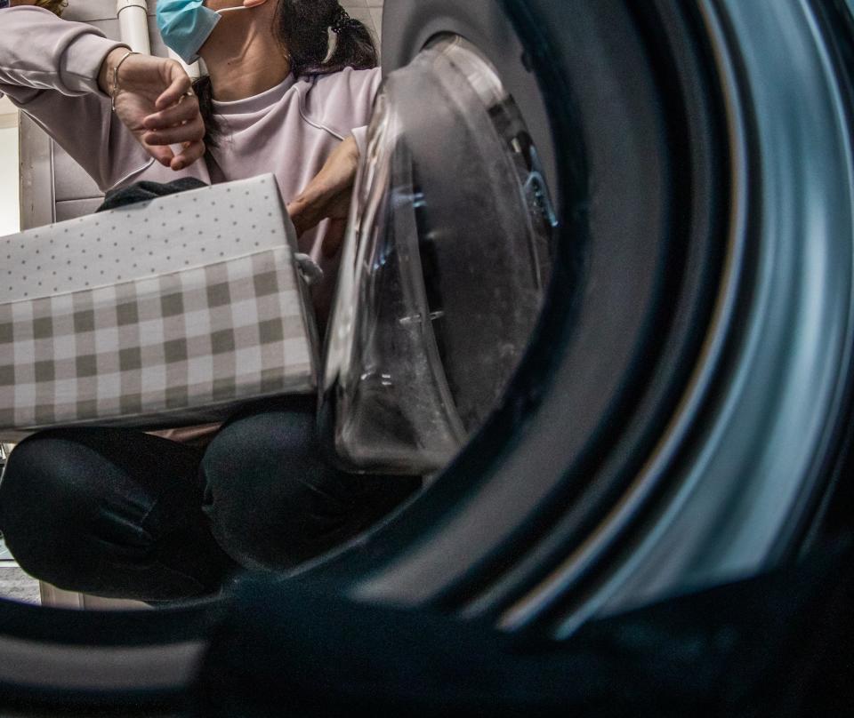 A man in scrubs and a woman wearing a mask, reaching for a checkered box inside a washing machine. A child looks on from the background