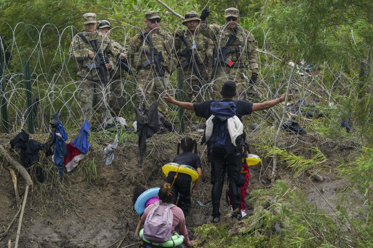 A migrant, surrounded by three small children wearing backpacks and toy flotation devices, holds his arms open wide as he looks at five Texas National Guards behind a fence of razor wire.