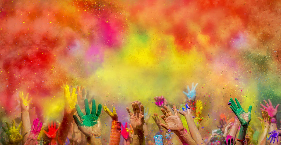 A vibrant scene of numerous hands reaching upward, covered in colored powders such as yellow, pink, blue, green, and red, likely at a Holi festival