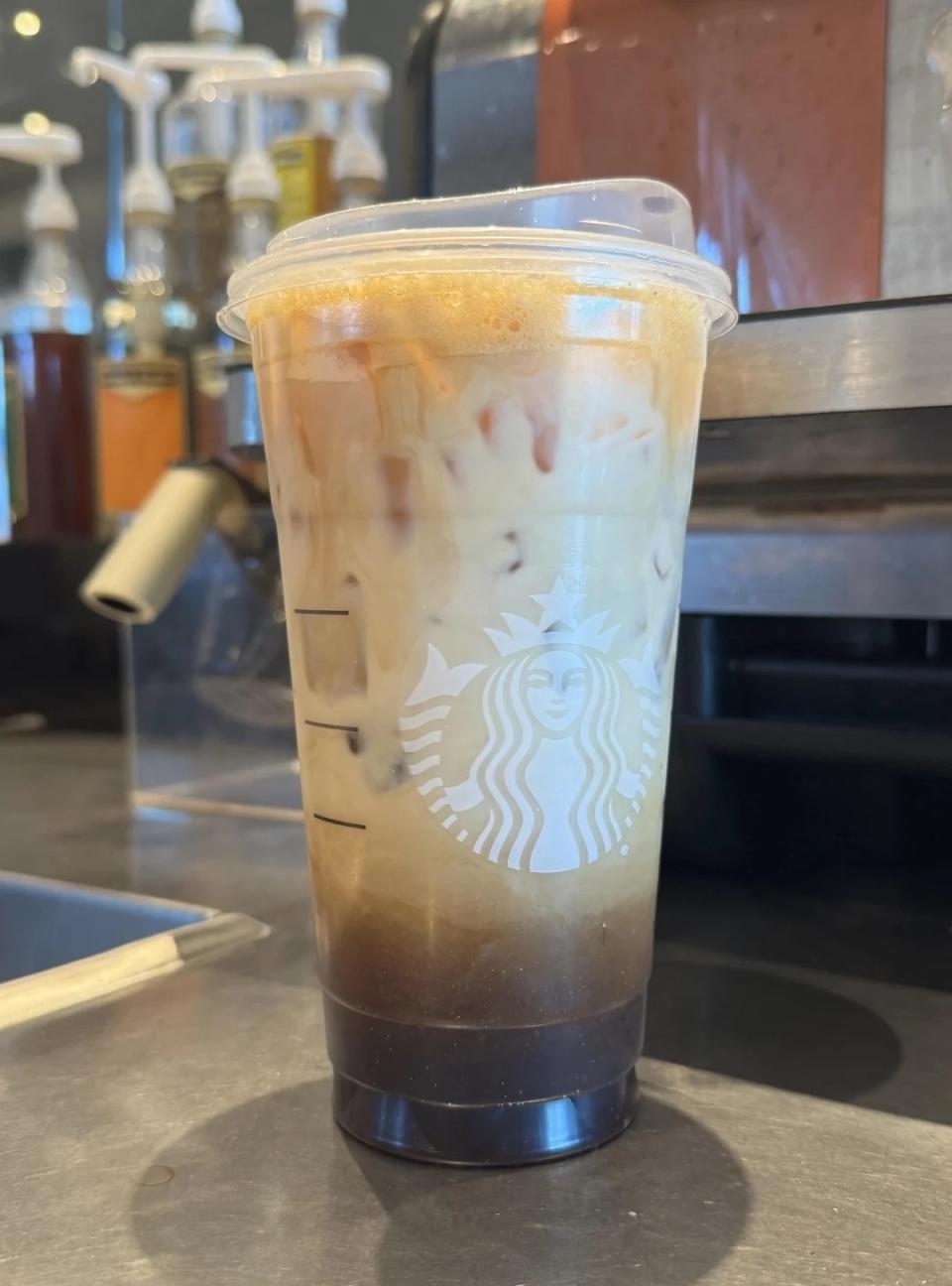 A Starbucks iced coffee beverage layered with milk and coffee, in a plastic cup with the Starbucks logo, placed on a countertop in a coffee shop