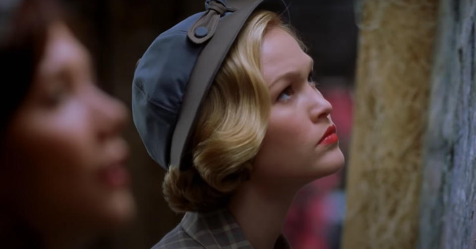 Julia Stiles, wearing a vintage-style hat, looks intently at something off-frame. Another person is blurred in the foreground.