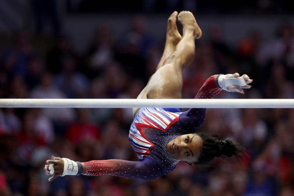 Simone Biles is captured mid-air in a gymnastics routine, performing on the uneven bars in a practice session. She is wearing a dynamic sparkling leotard