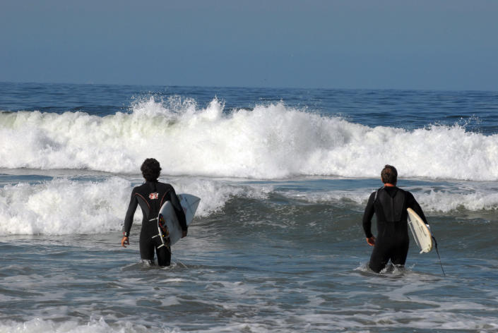 دو موج سوار در آب ایستاده اند و امواج را تماشا می کنند.