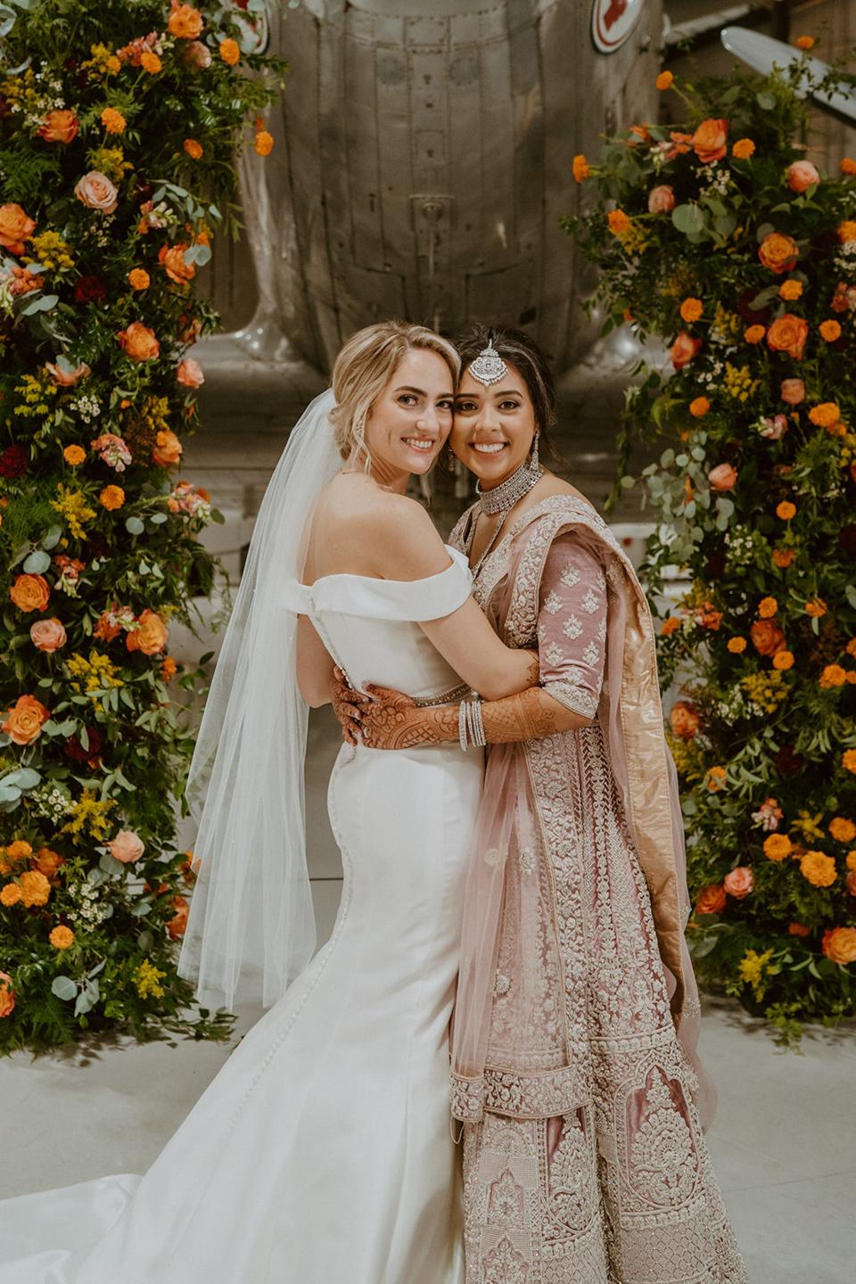 Two brides smile and hug in front of a floral archway.