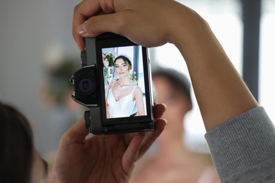 Bride in a white dress holding a bouquet, captured on a camera's screen, others blurred in background