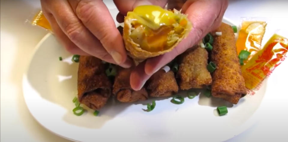 A person's hands holding a half-eaten egg roll above a plate with more egg rolls and sauce packets