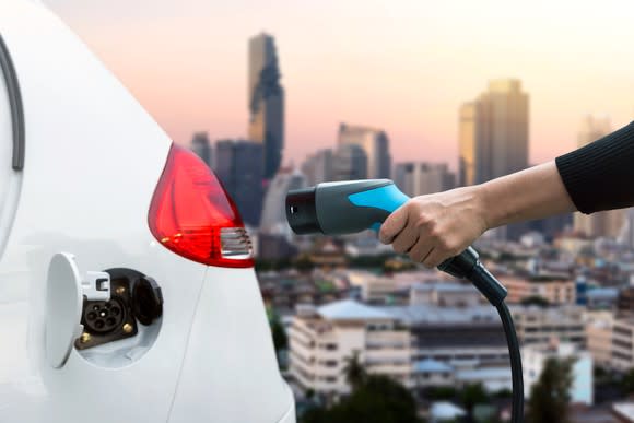 Person's hand holding an EV charger and about to charge (or just finished charging) his or her white vehicle. A city in background.