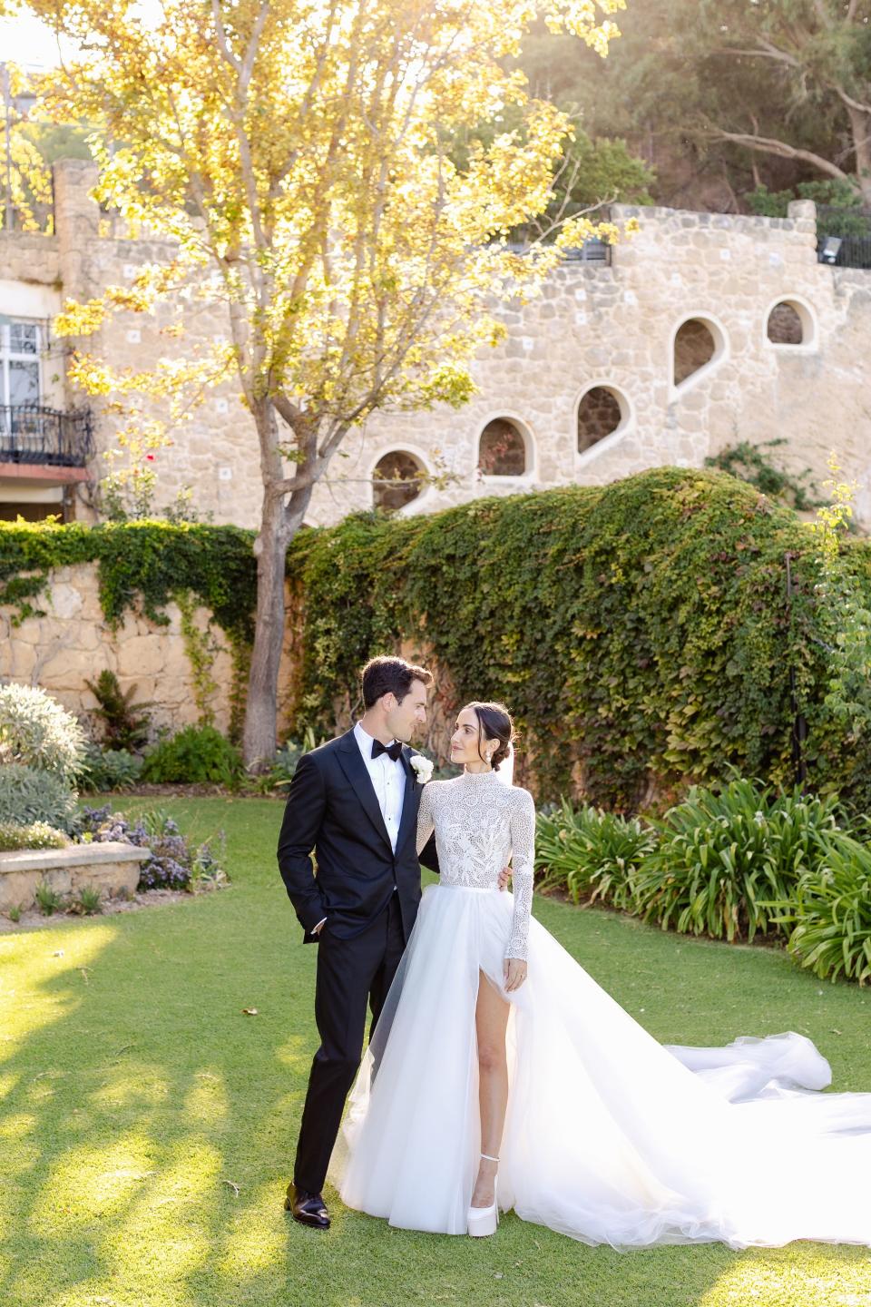 A bride and groom pose in a field in their wedding attire.