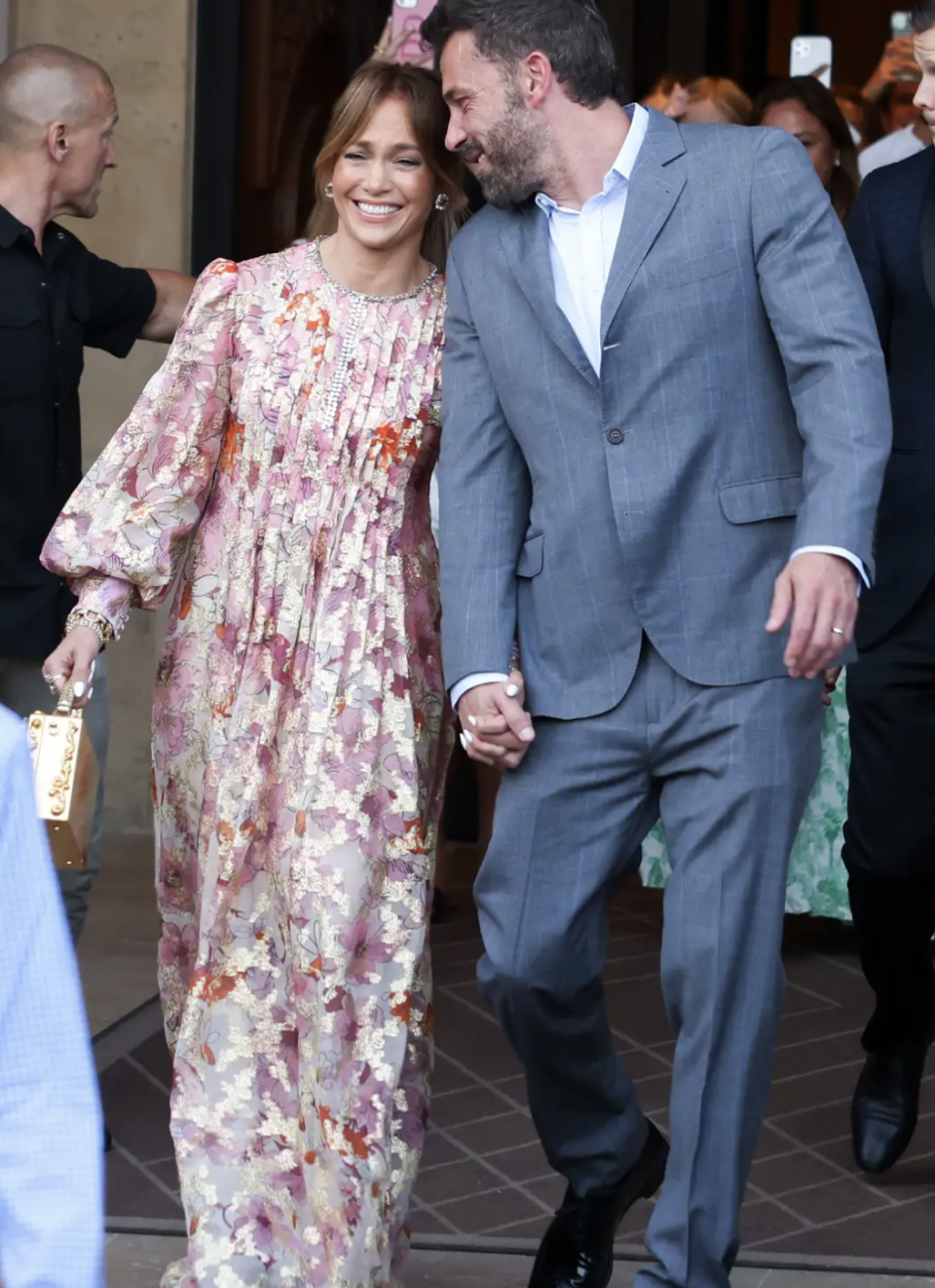 Jlo and Ben Affleck holding hands and smiling with Jen in floral dress and Ben in charcoal suit