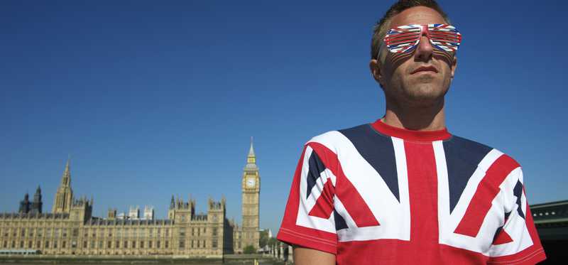 A young man wearing a British flag shirt and sunglasses, with Parliament in the background.