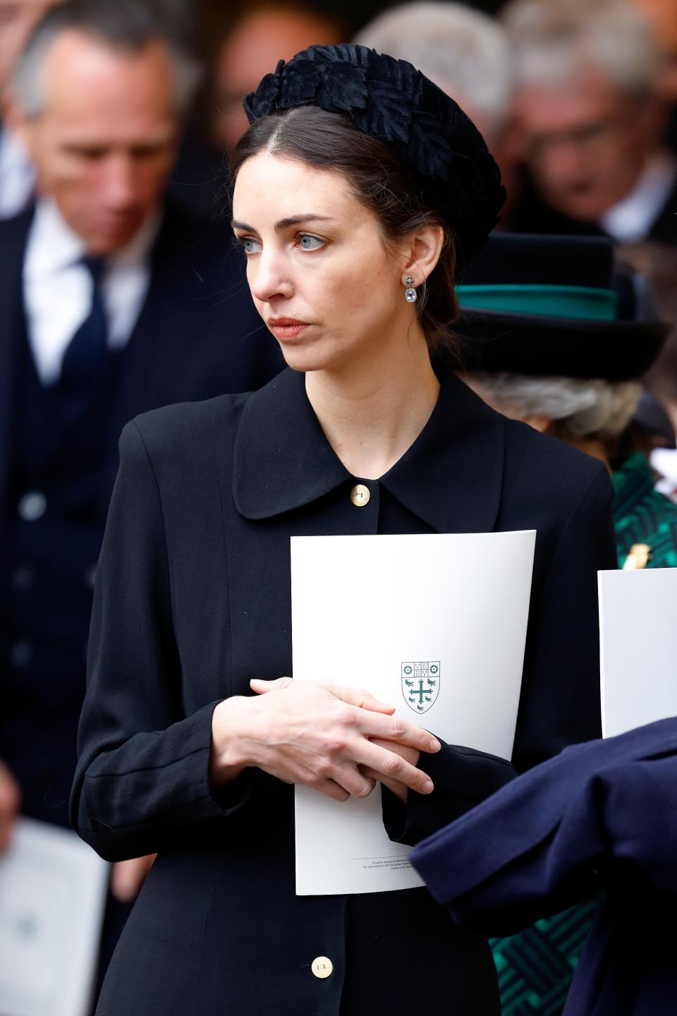 Rose Hanbruy in a solemn gathering wearing a black coat and hat, holding a service booklet