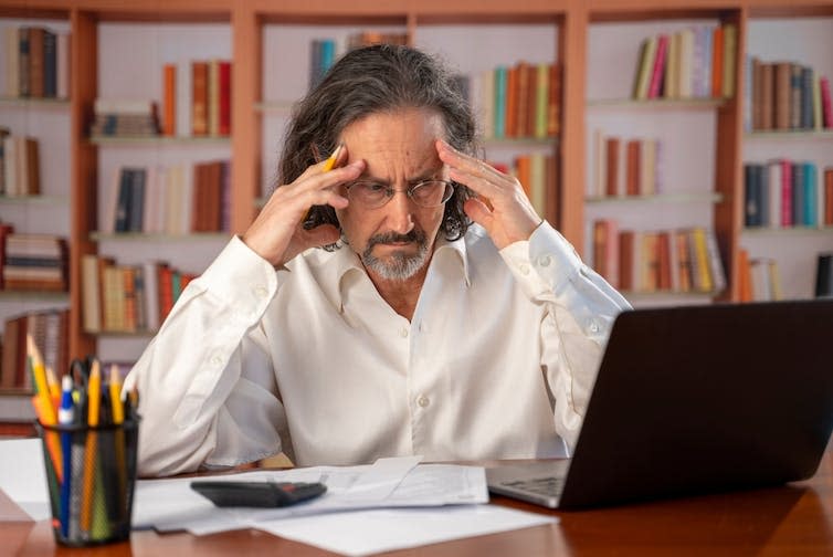 A frustrated manager in front of a laptop at a desk; man with long hair and beard at a desk, hands to head.