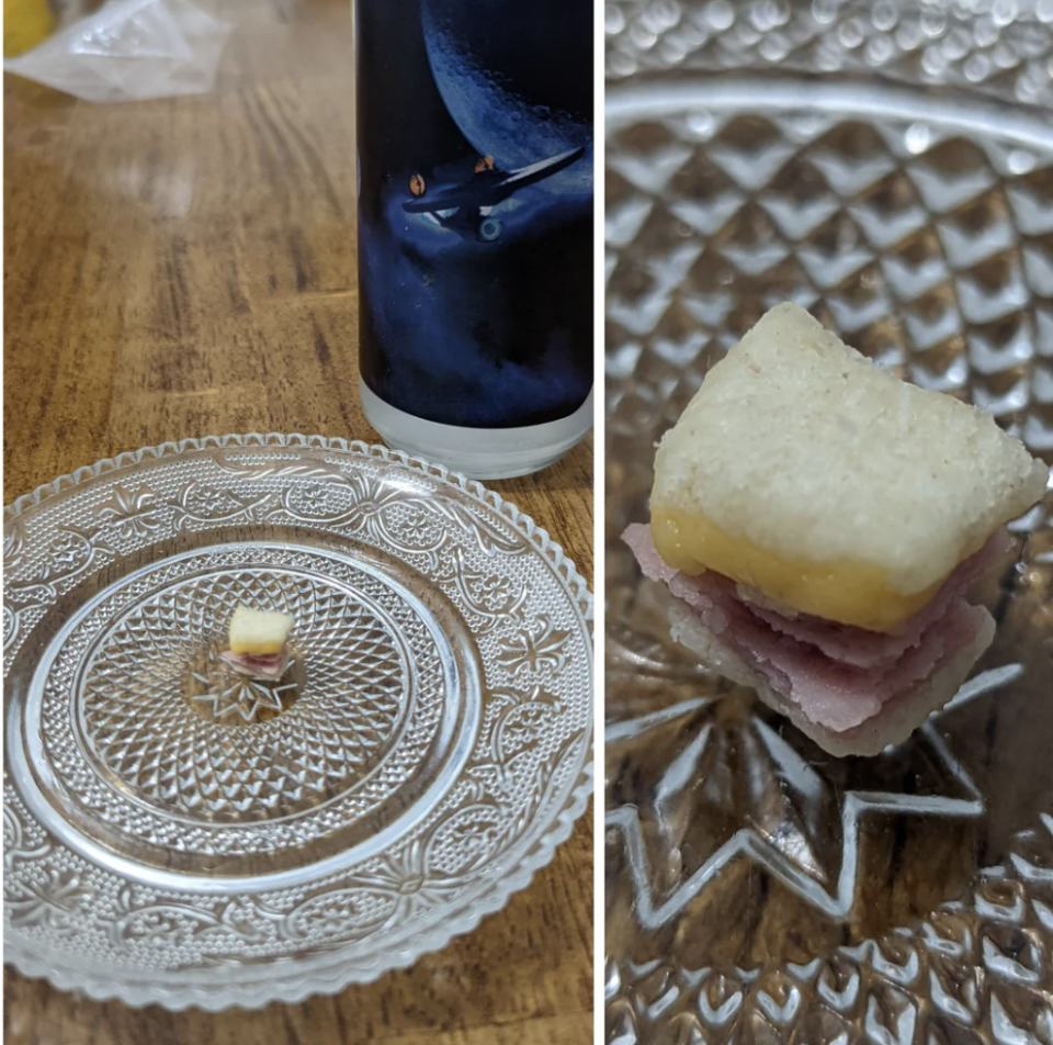 Close-up of a small, sandwich-like snack on a decorative clear plate. The snack has layers of bread, meat, and a yellow topping. A decorative candle is visible