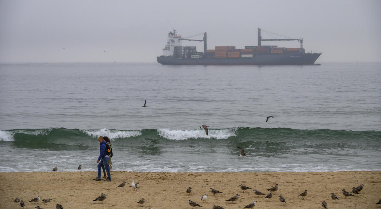 MATOSINHOS, PORTUGAL - SEPTEMBER 19: A couple walks surrounded by seagulls in Matosinhos beach while Container ship Ludwig Schulte is seen through the afternoon fog while sailing to the port of Leixoes on September 19, 2019 in Matosinhos, Portugal. The Port of Leixões is the largest port infrastructure in the Northern Region of Portugal and one of the most important in the Country. (Photo by Horacio Villalobos#Corbis/Corbis via Getty Images)