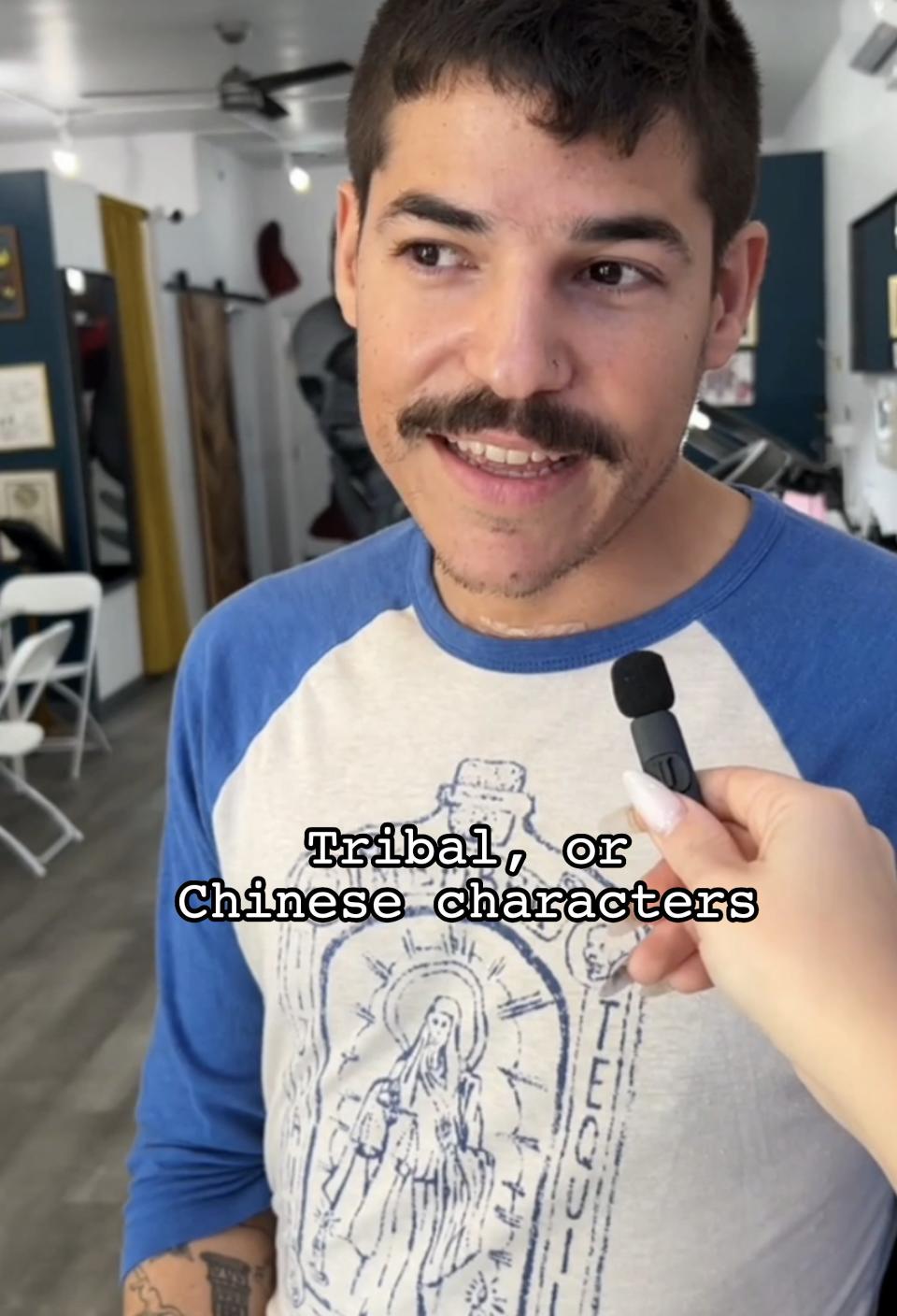 Man with mustache interviewed, wearing graphic tee, indoors with blurred background