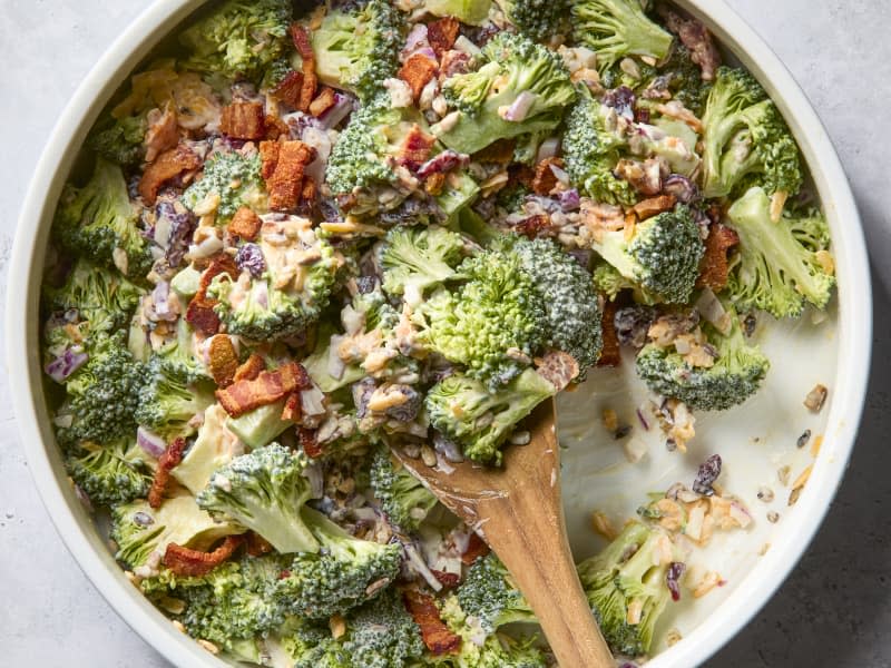 overhead shot of a mixed up and dressed broccoli salad, with some missing out of the left side of the bowl and a wooden spoon resting in the bowl.