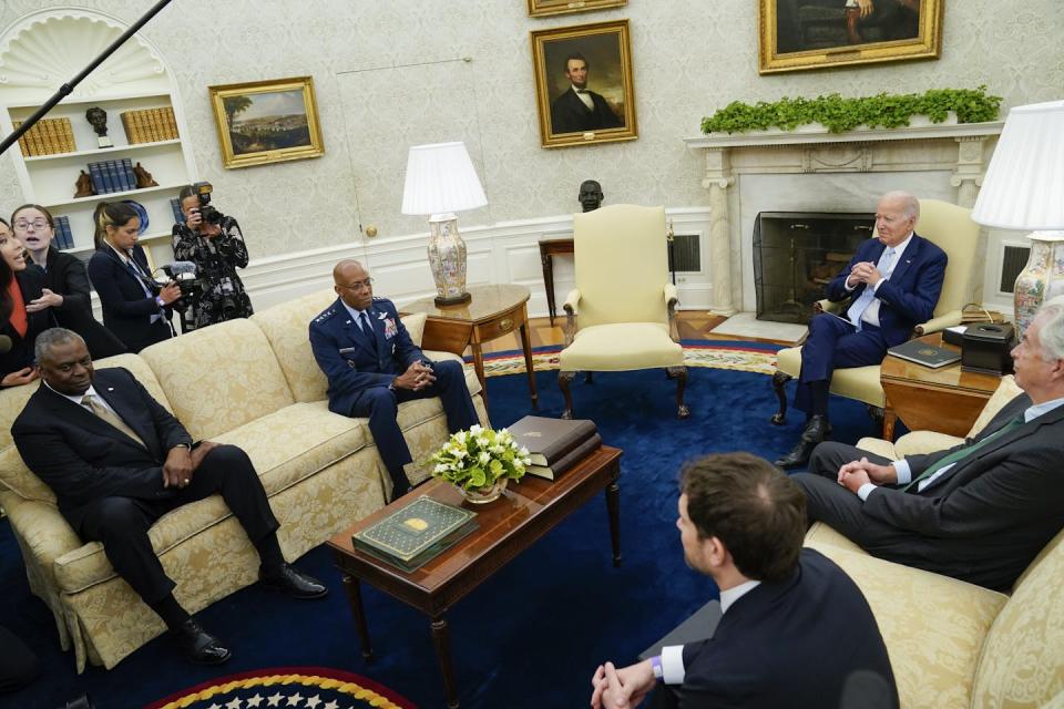 President Biden sits in a chair with other men in suits on couches in an oval room at the White House
