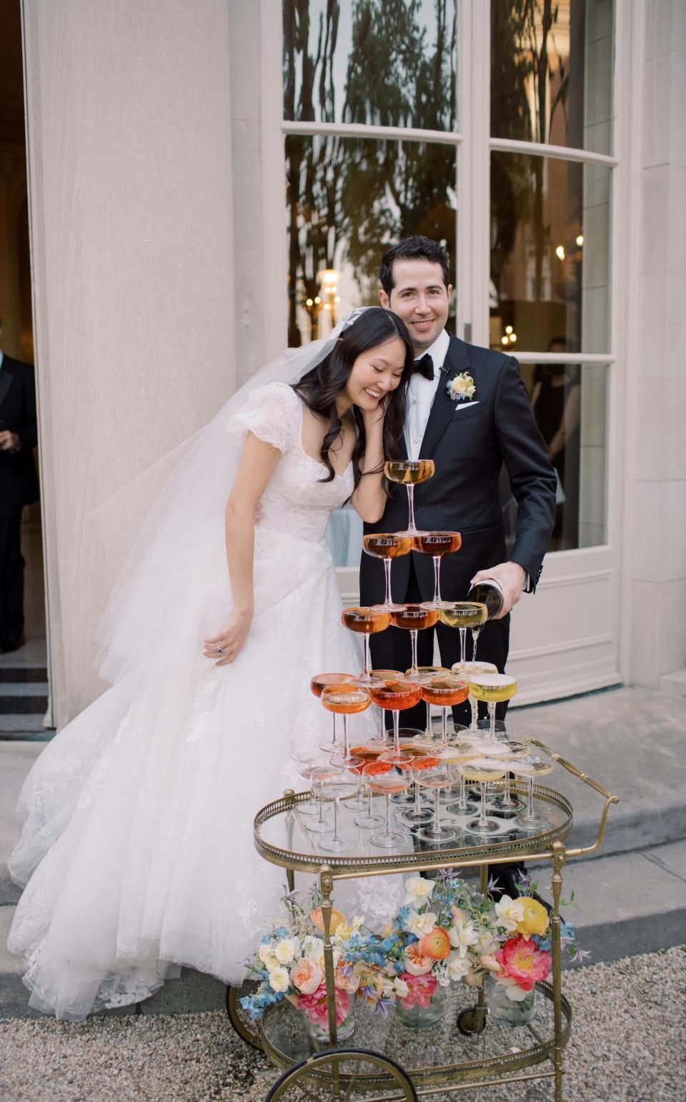 A bride and groom lean together in front of a champagne tower.