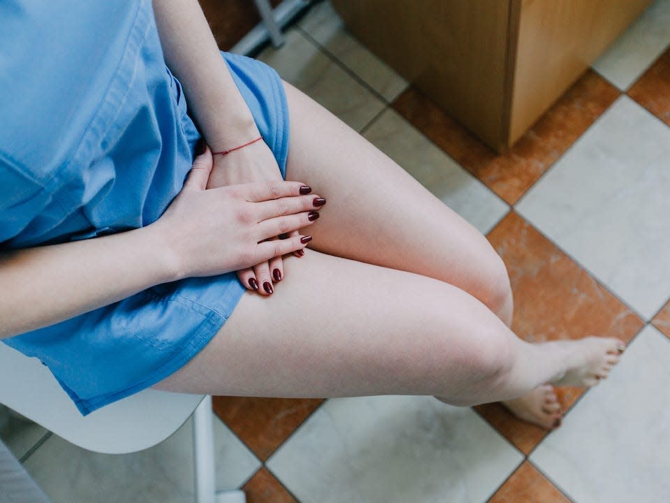 A woman wearing a blue medical gown crosses her legs and covers her crotch with her hands.